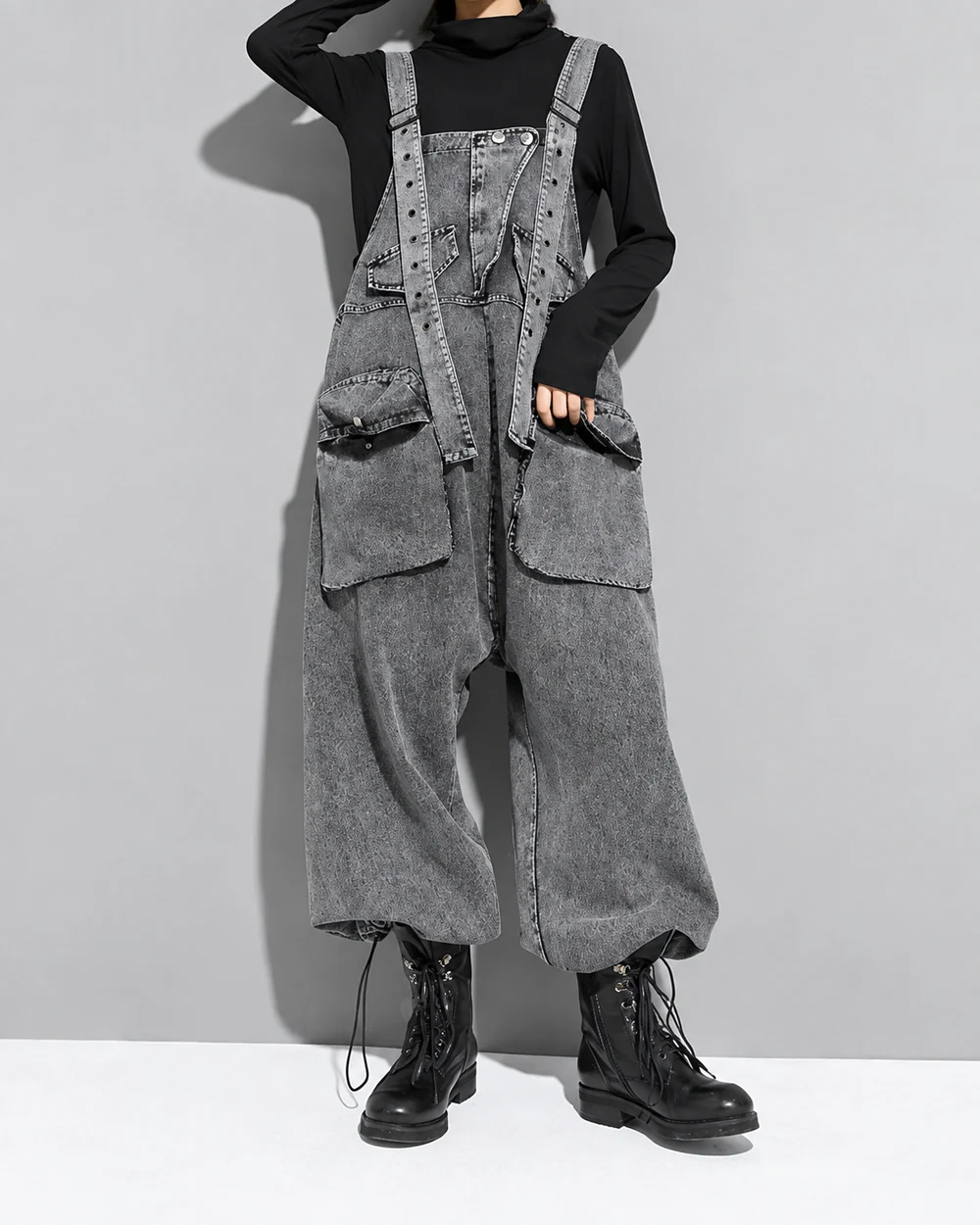 Faceless model wearing oversized gray-wash denim overalls with large utility pockets and balloon legs